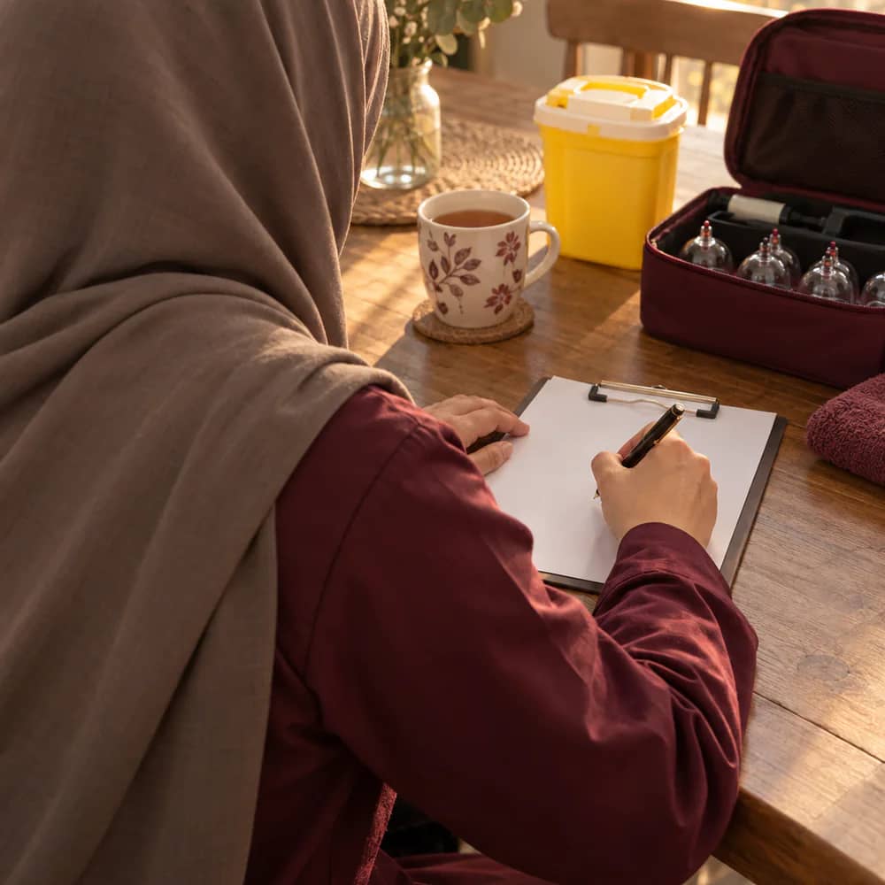 A hijabi practitioner, viewed from behind, writing notes beside a cup of tea after a home visit